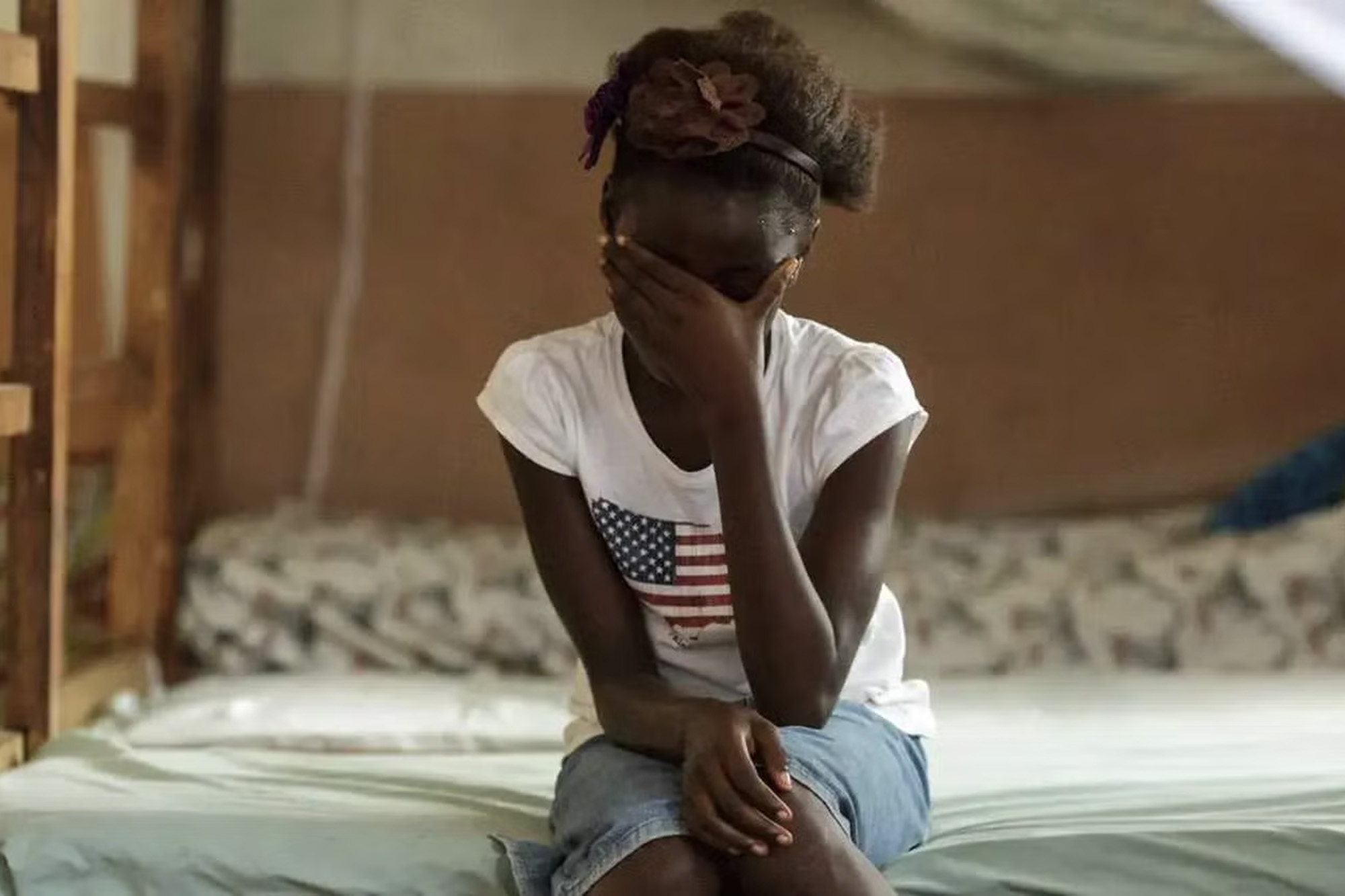 A woman sits on a bed and covers her face with her hand.