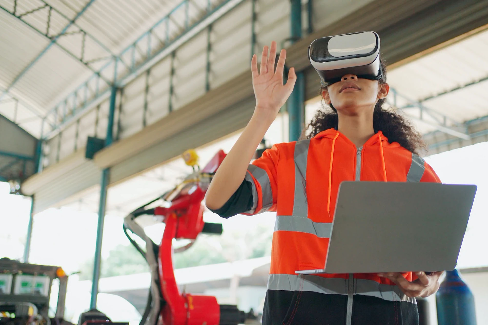 Woman wearing a virtual reality headset, holding a laptop in her left hand and drawing an imaginary space with her right hand.