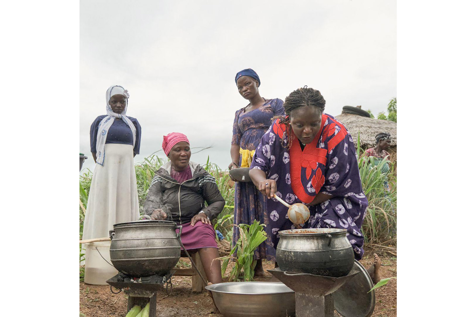 Four women cooking outdoors, with three standing and one sitting. The person sitting is stirring a pot on a stove, while another person is also attending to a pot.