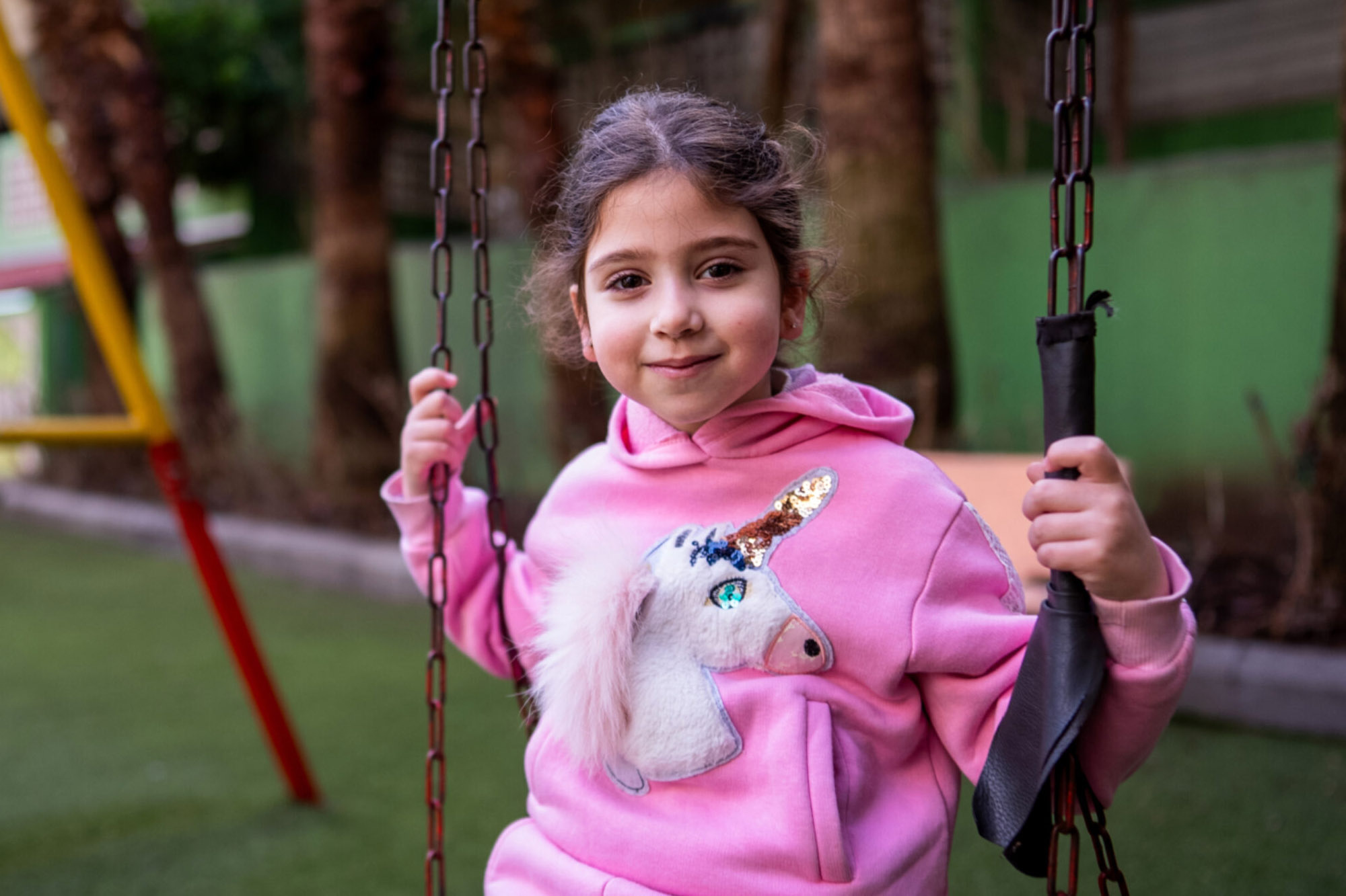 A child wearing a pink hoodie is sitting on a swing in an outdoor playground.