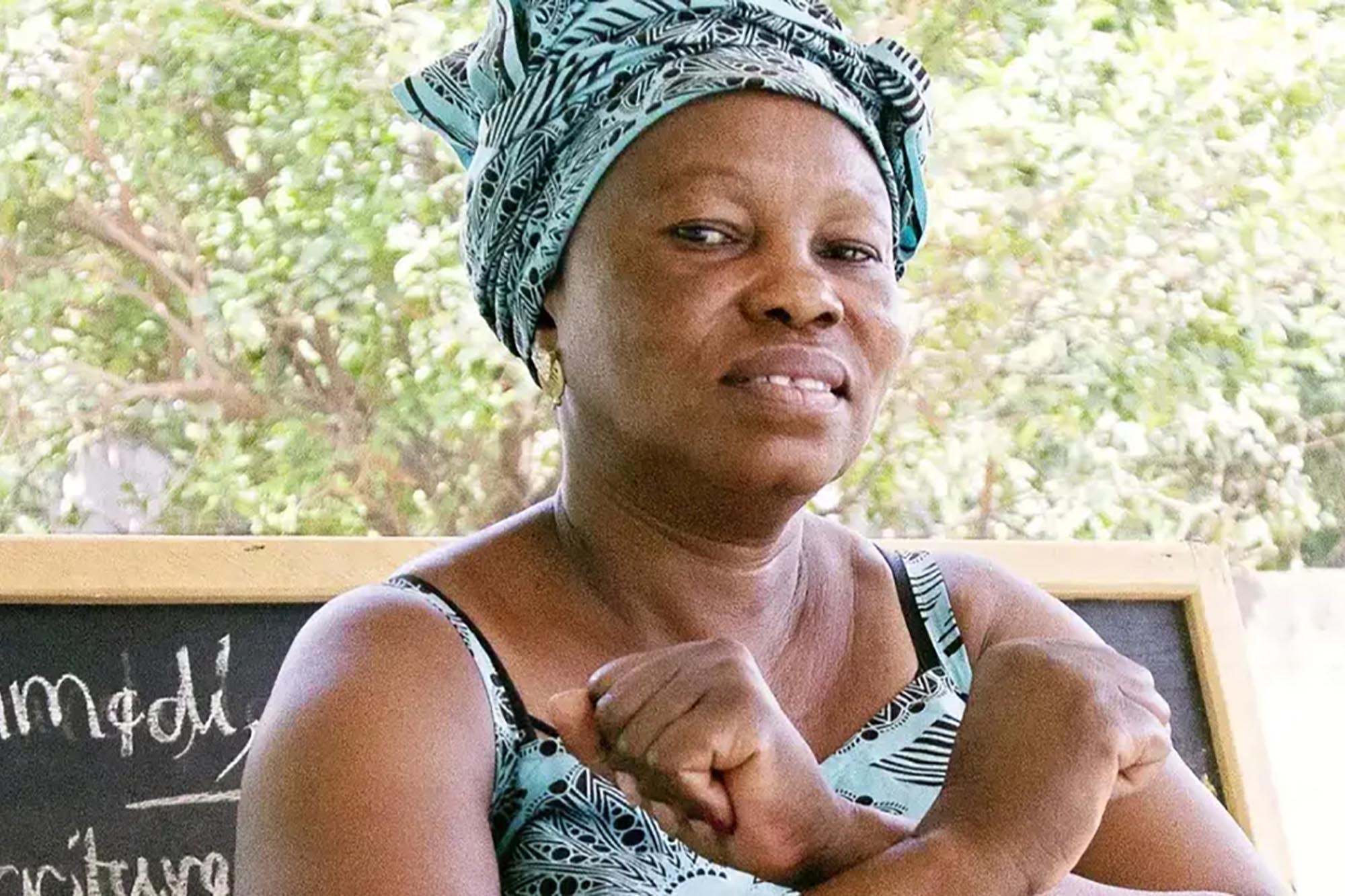 A woman standing confidently in front of a blackboard, dressed in patterned clothing with a matching headwrap.