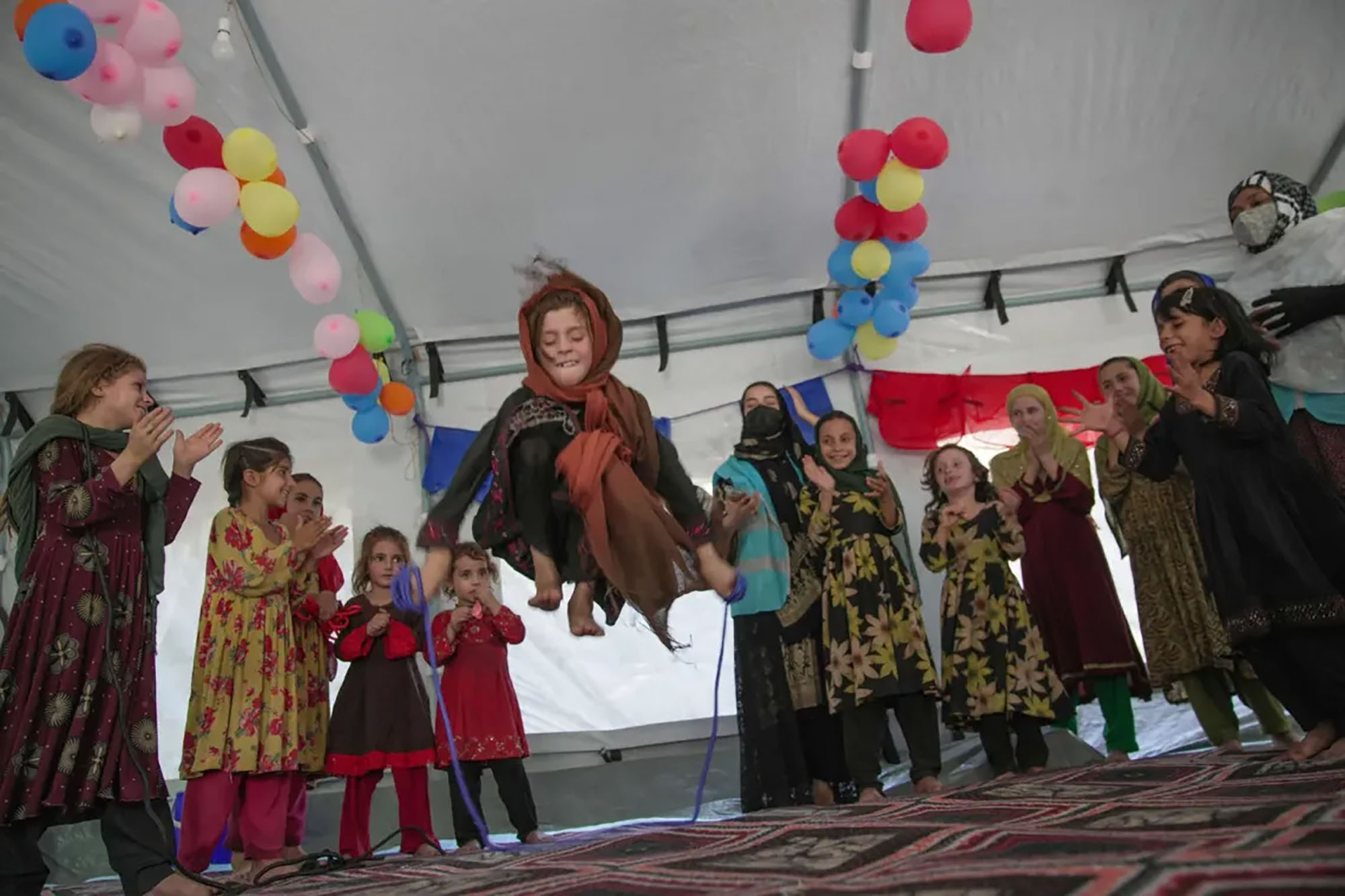A group of children dressed in vibrant, traditionally patterned clothing are gathered in a circle inside a tent decorated with colourful balloons hanging from the ceiling.