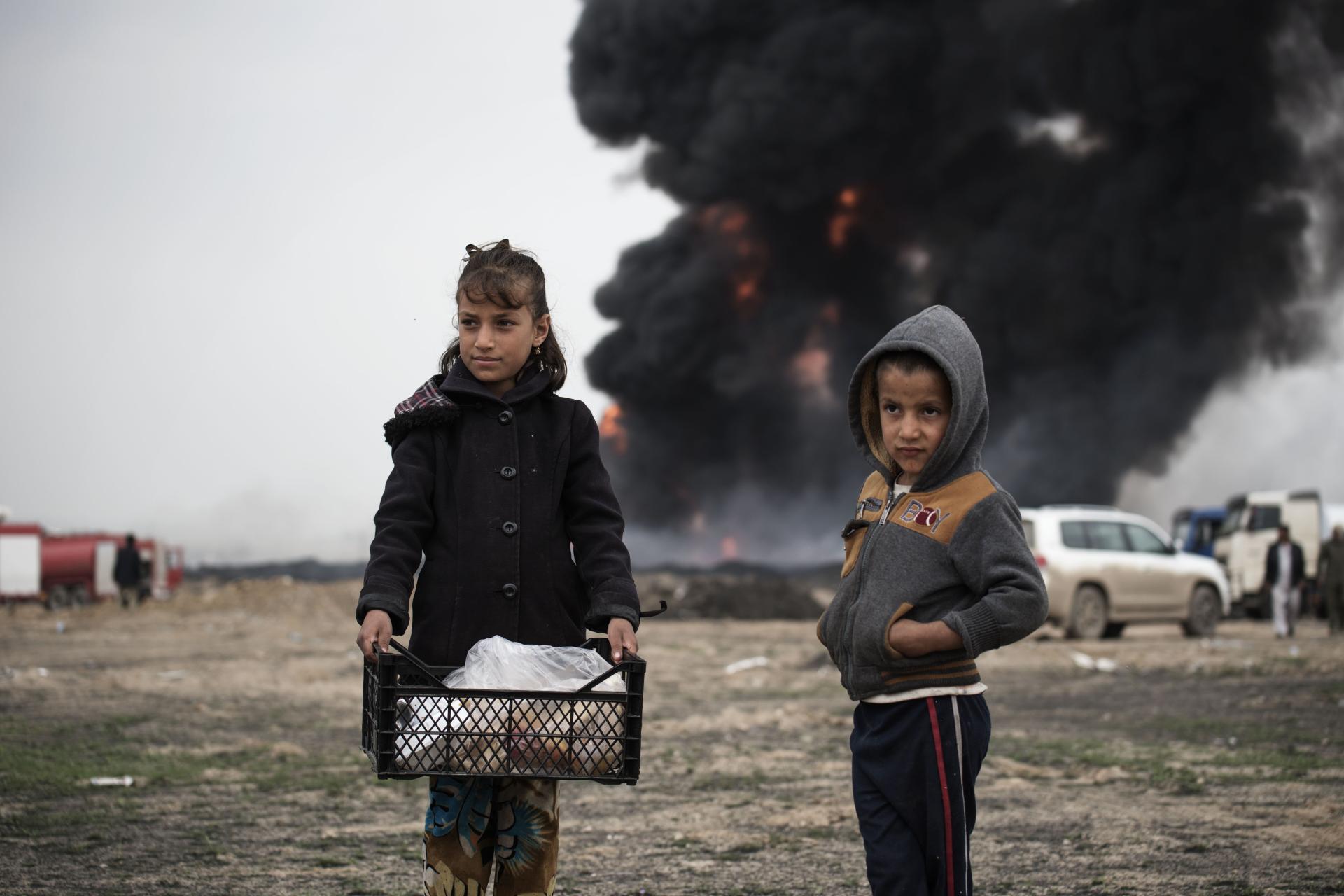 a girl and a boy look out with dismay as a fire burns behind them in a scence of war