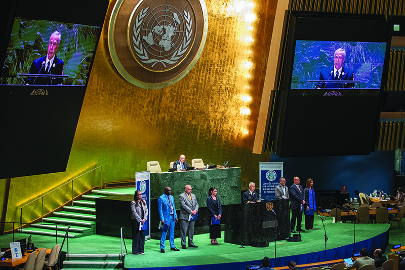 View of the General Assembly room. A green podium stands behind a black one. A man is speaking at the podium, while three people stand to his left and three to his right. Another man is seated at the green lectern behind them.