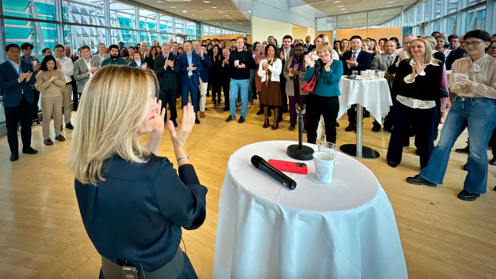 Celeste stands in a large room facing a large group of people as they all smile and some clap their hands
