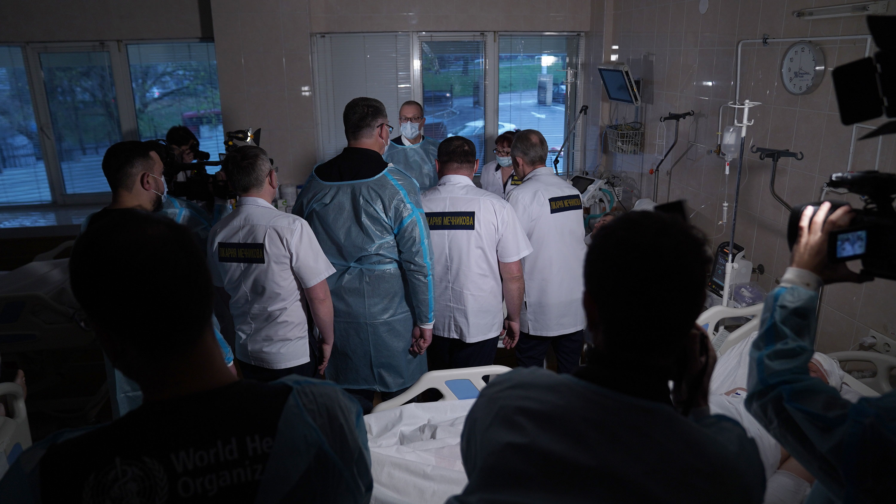 Hans next to a hospital bed with the windows behind him and in front of him is a throng of people with a camera-person at the forefront. All are wearing facemasks.