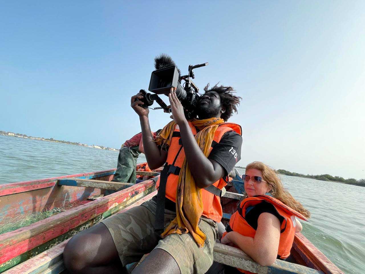 Carmen sits in a motorboat while a man, seated ahead of her, holds a large camera on his shoulder and films the scene ahead
