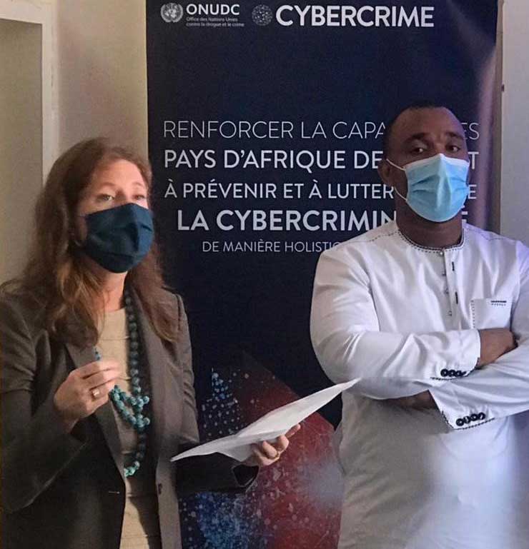 Carmen stands in front of a large poster, holding a paper in her hand, delivering a speech whilst wearing a COVID face-mask. A man, also wearing a mask, stands beside her.
