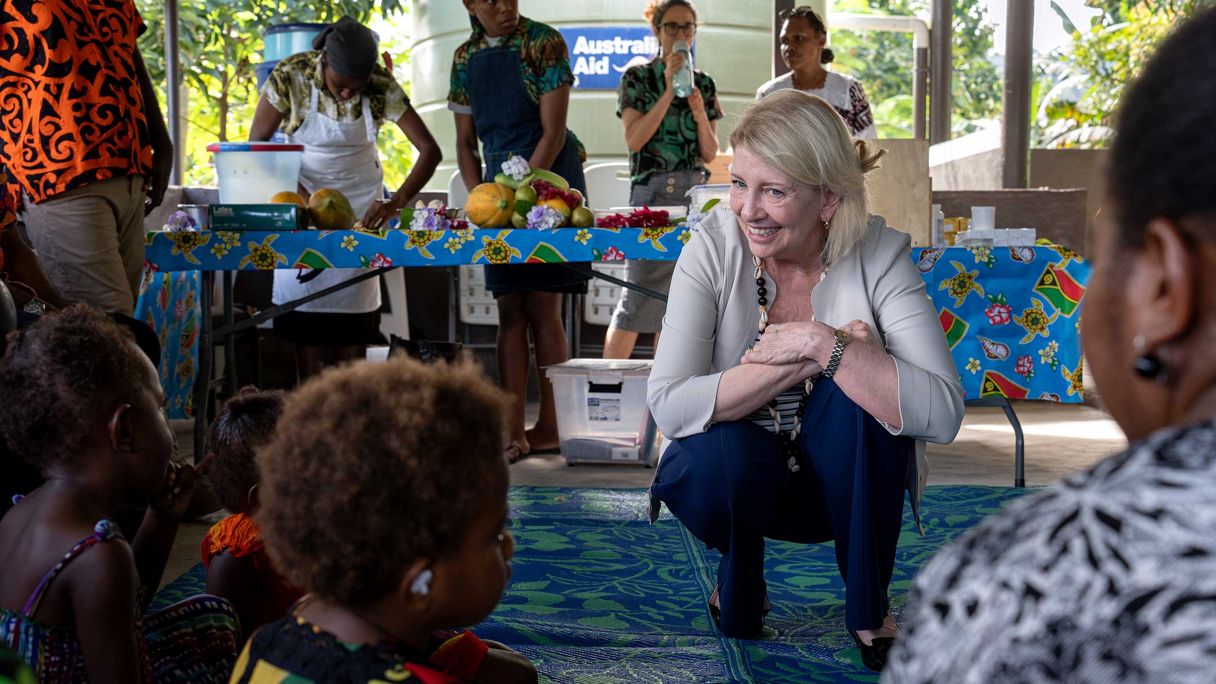 Catherine is crouching and looking at young girls while people in the background prepare food for distribution
