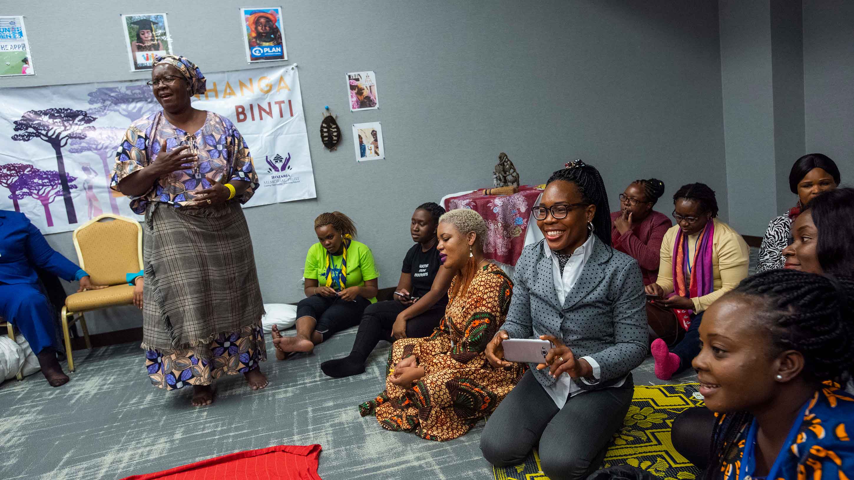 Nyaradzayi stands in a room full of people sitting on the floor next to her