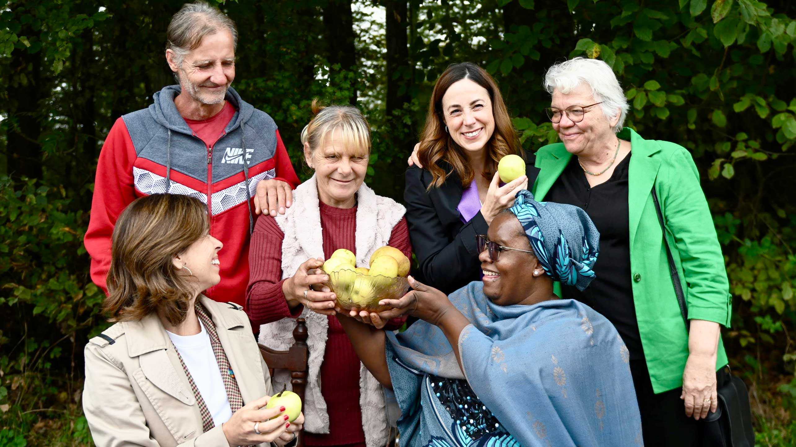 In a group photo with 5 people, Nyaradzayi hands over a bowl of yellow apples and pears to the person standing behind her