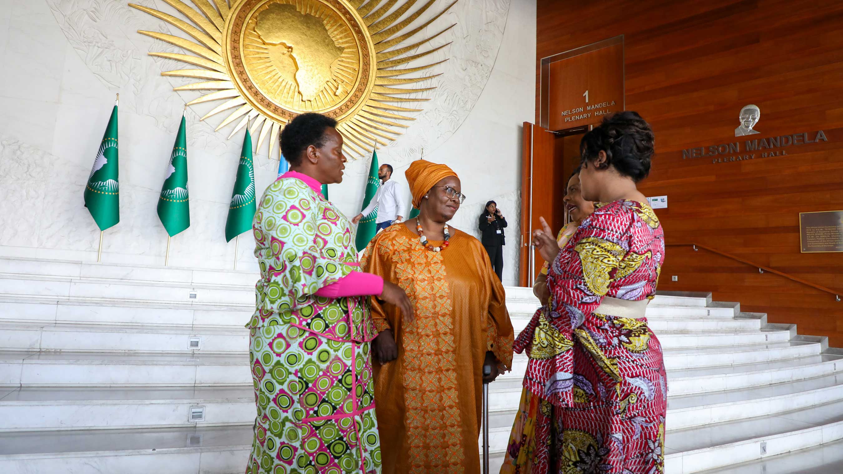 Nyaradzayi is speaking with 2 women at a large staircase with a golden radiating circle relief-sculpture of the African continent seen hanging behind all