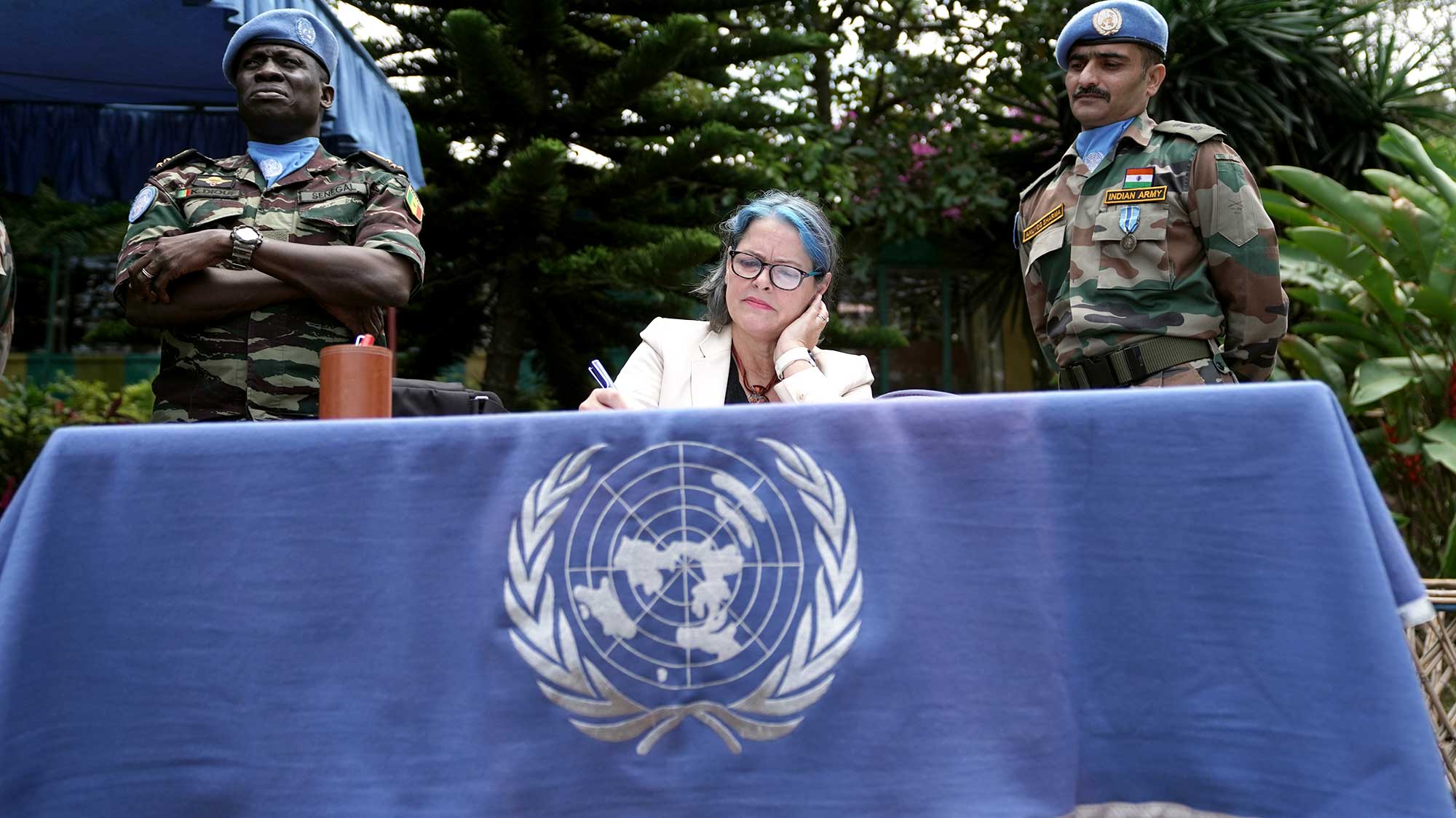 Vivian sittin g at a table draped with the UN flag and she is writing into a book with two UN troops standing behind her