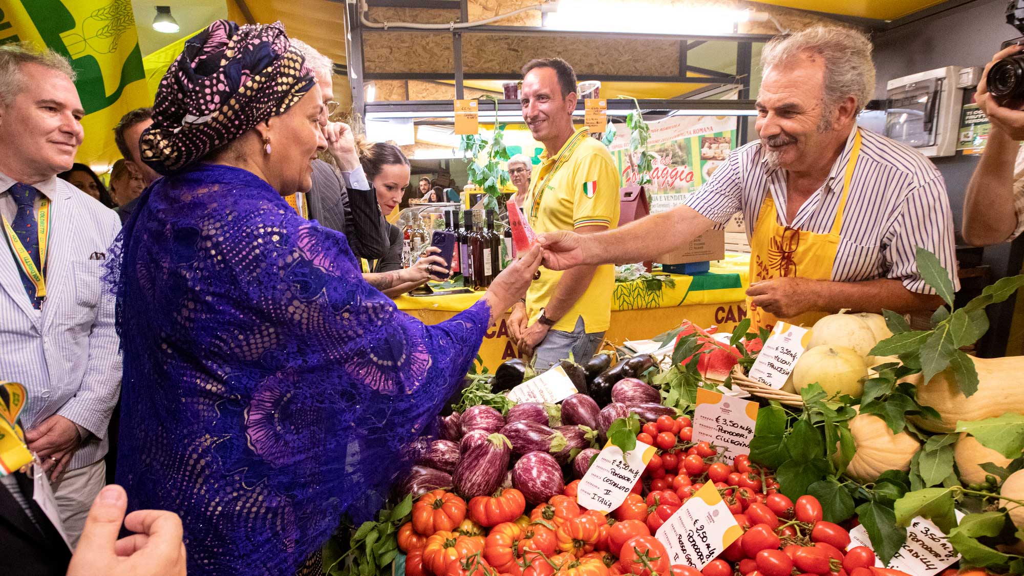 Amina takes a watermelon wedge from a farmer who is handing it to her at a farmers market stand