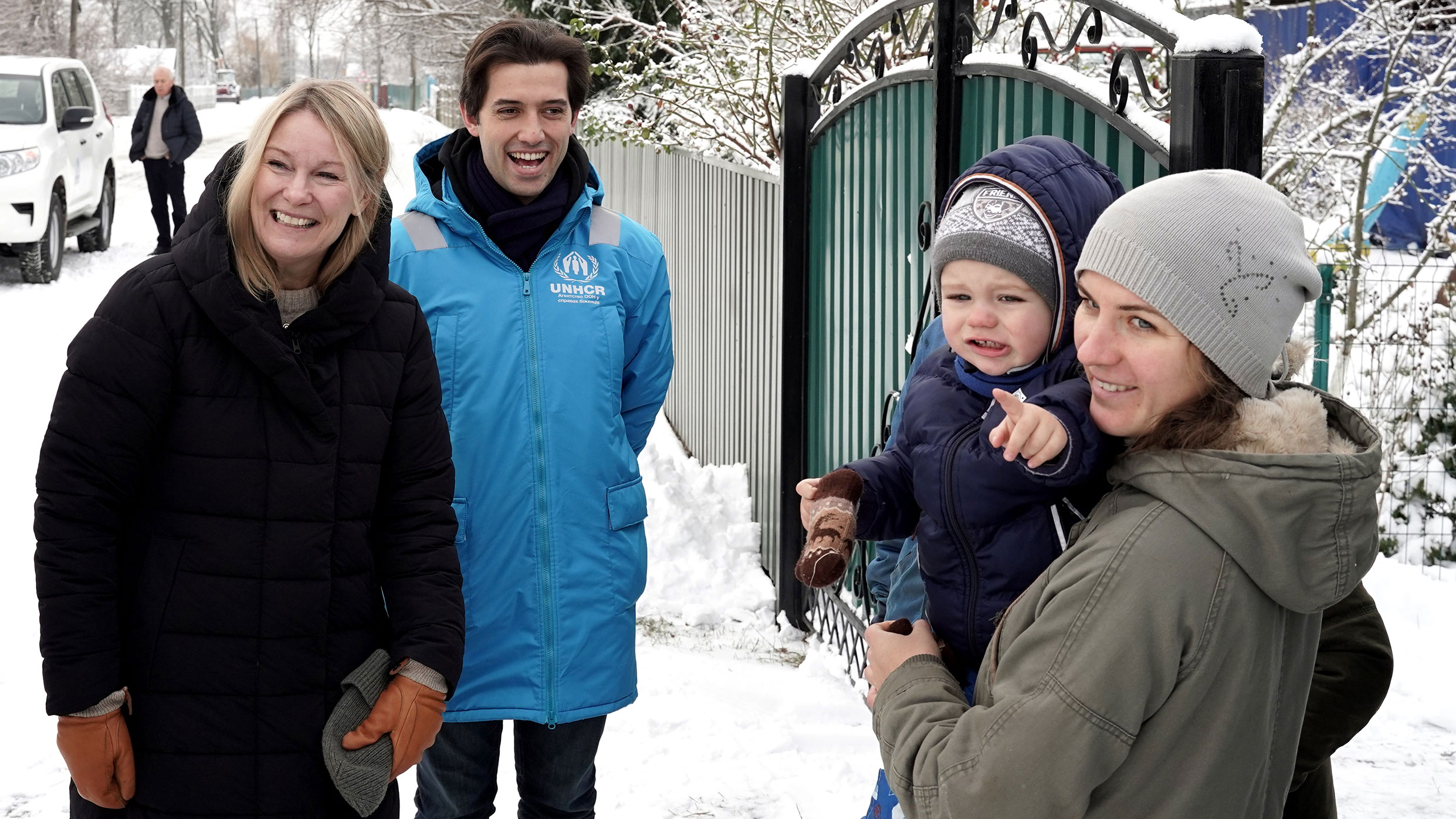 Karolina stands in the snowy outdoors next to a woman carrying a child and a young man in a blue UNHCR vest
