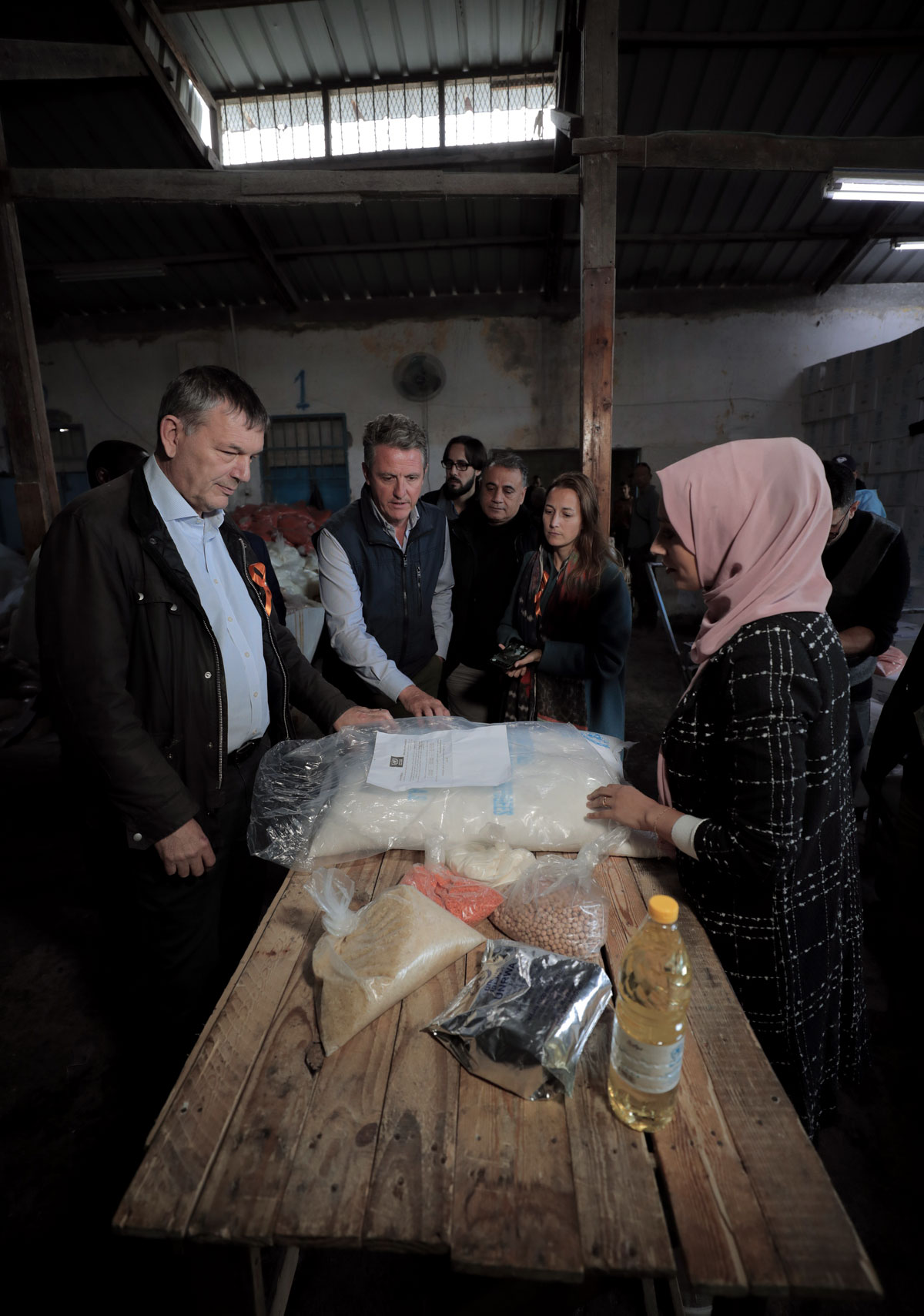 Among a crowd, Lazzarini listens to a woman wearing a veil. They are inside a warehouse distribution center and some supplies are on display on a table.