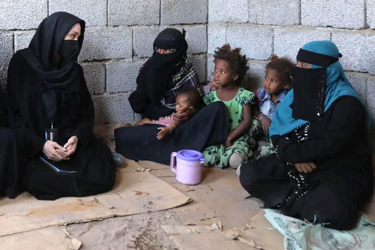 Angelina Jolie, wearing a hijab, sits on the floor next to women and girls.