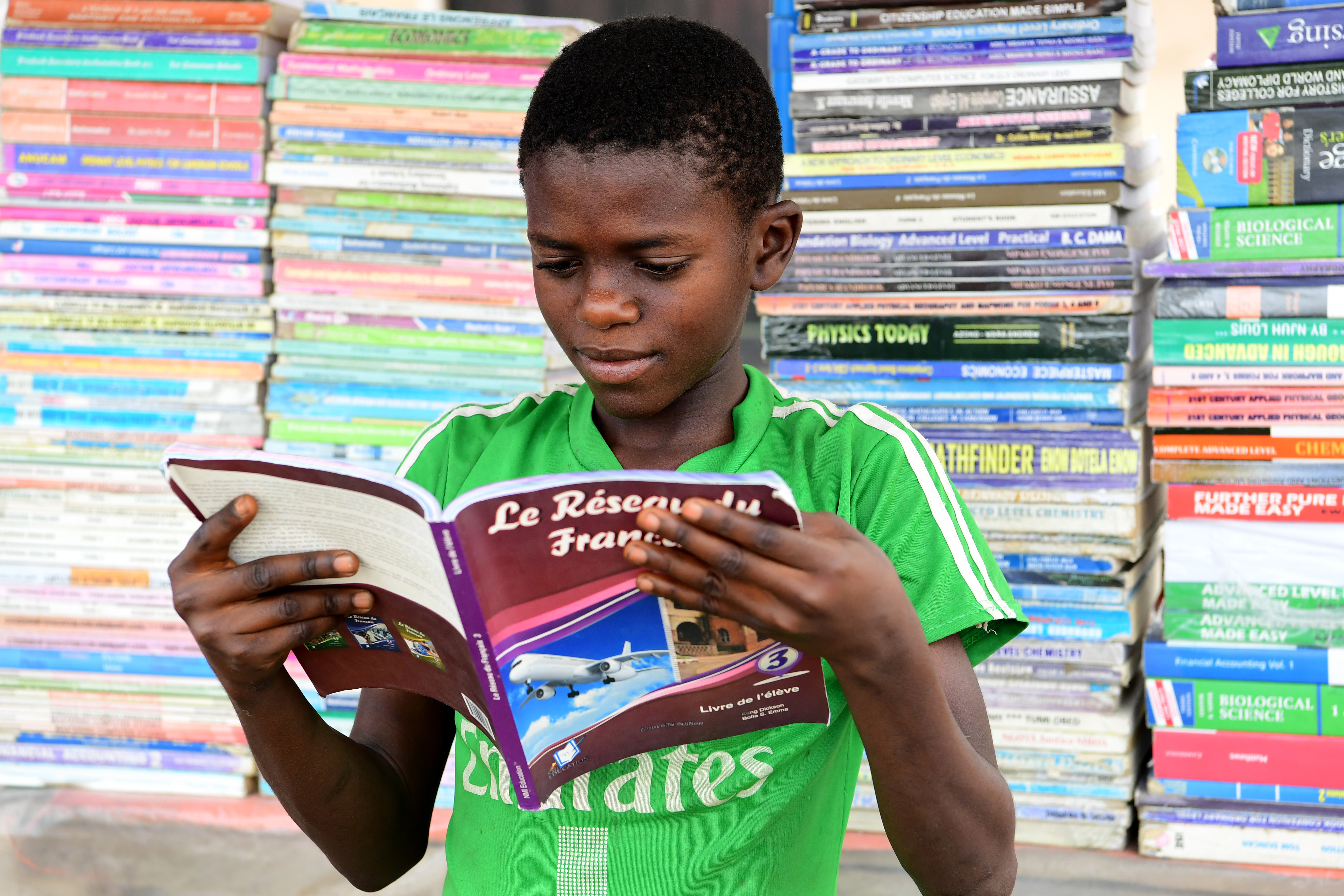 A boy reads a book in front of stacks of other books. 