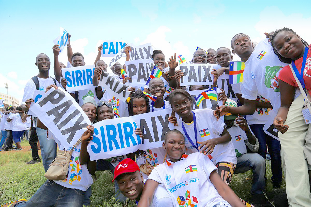 A group of joyful youth come together playfully holding signs that read peace.