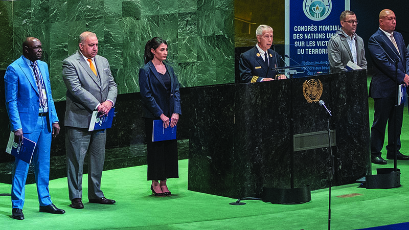 View of the General Meeting room. A green podium stands behind a black one. A man is speaking at the podium, while three people stand to his left and three to his right. Another man is seated at the green podium behind them.