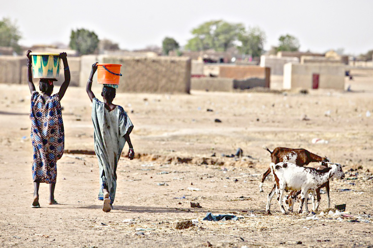 Two kids carrying water in buckets with a dryland as a background.