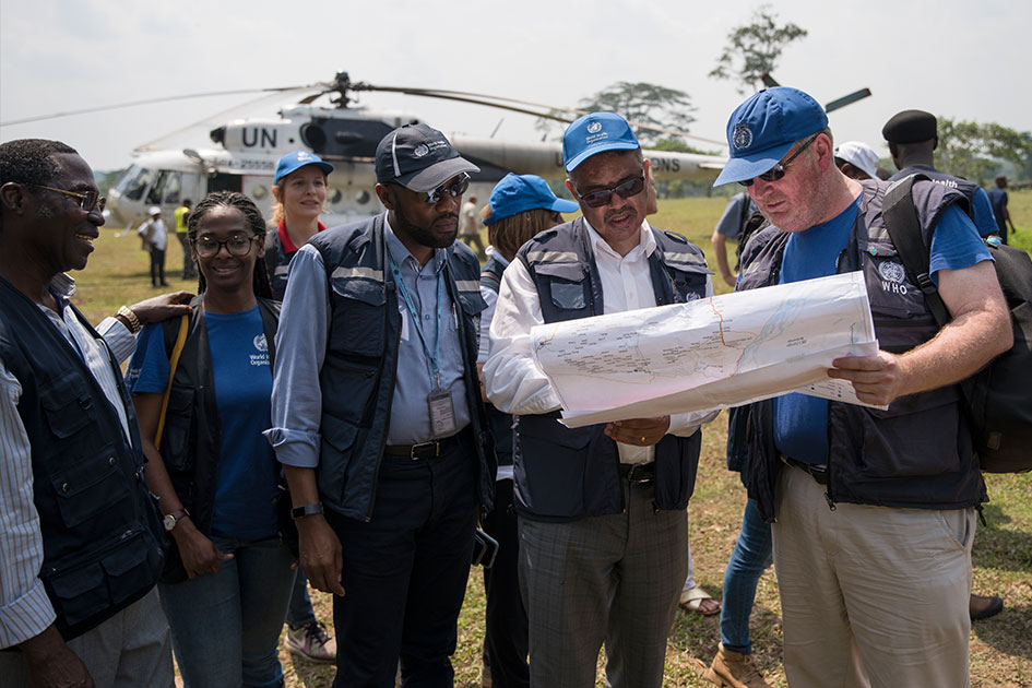 Dr. Ryan and Dr. Ghebreyesus are looking at a map. They are surrounded by their team. A helicopter is seen in the background.