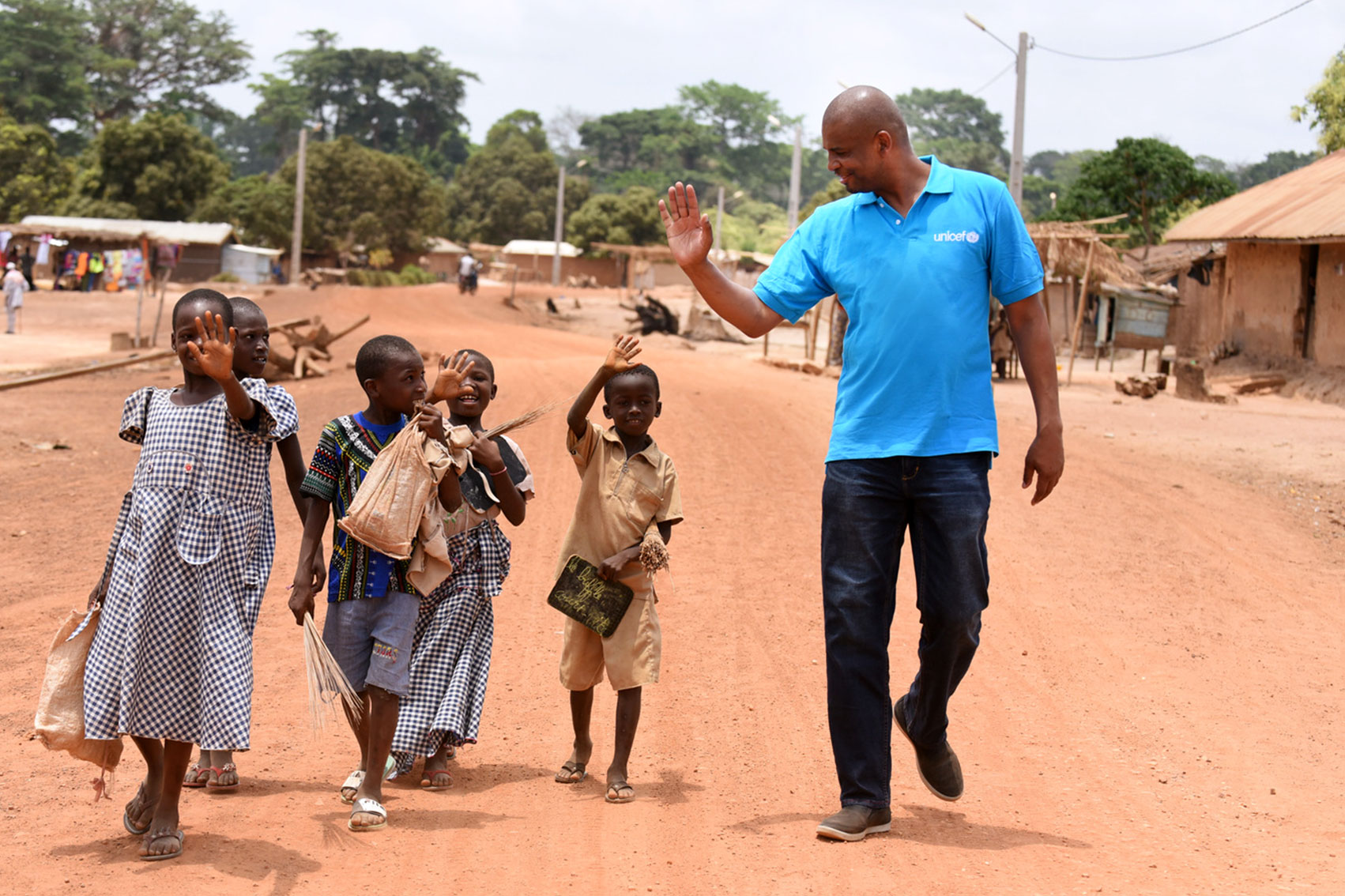 Aboubacar walking along a dirt road and children are walking on his right side. They are all waving at each other.
