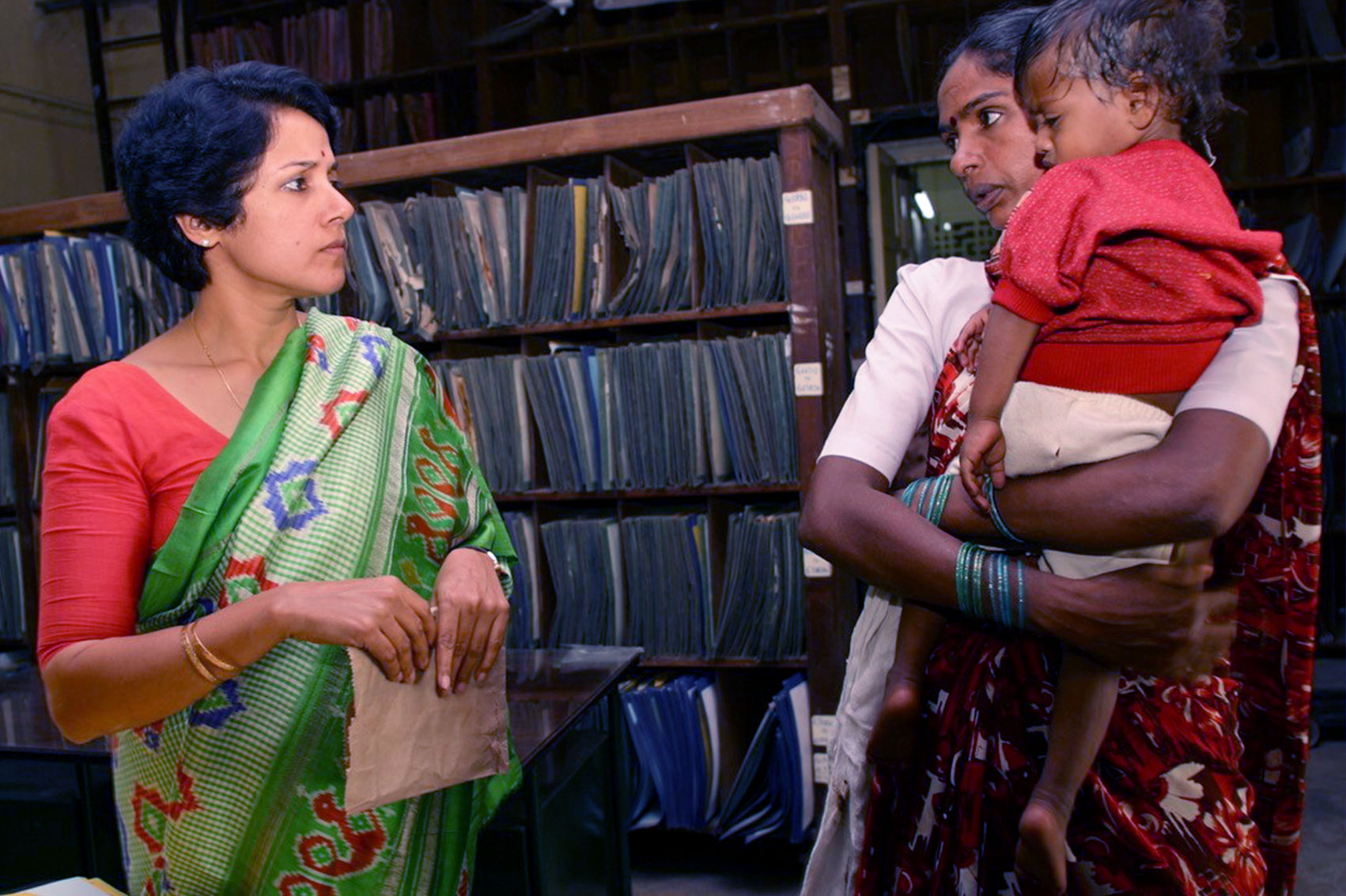 Dr. Swaminathan stands in an office with a mother holding a child next to her.
