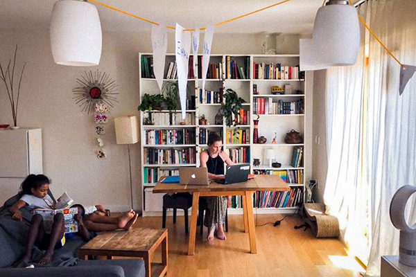 Woman works from two laptops with two children on a couch next to her desk.