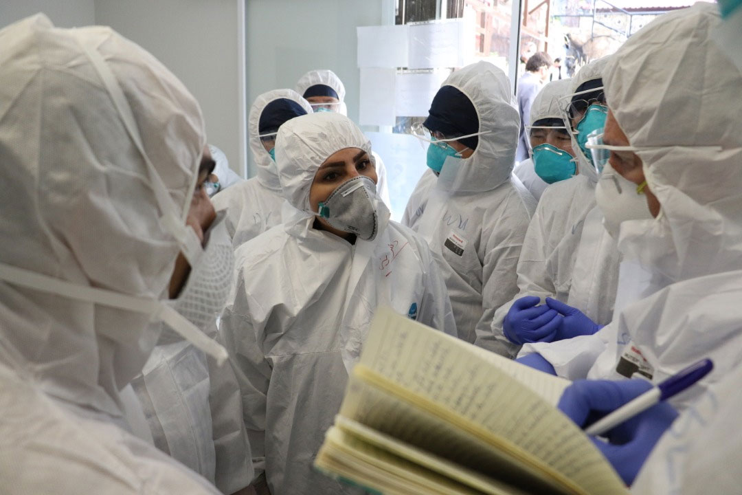 WHO and public health experts are deep in discussion whilst one writes notes. They are dressed in head-to-toe protective gear including face masks.