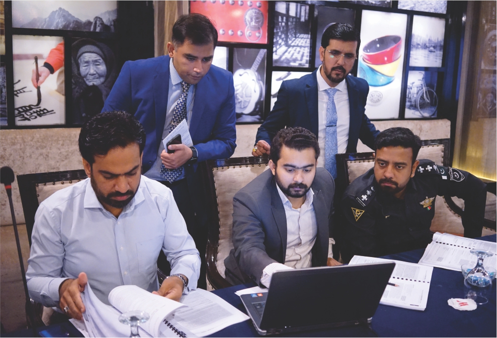two men stand behind three seated men and they look over their shoulder whilst working on files and a laptop