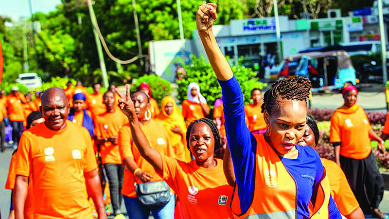 People march down the street. They are all wearing orange clothing. Two girls in the foreground have their arms raised.