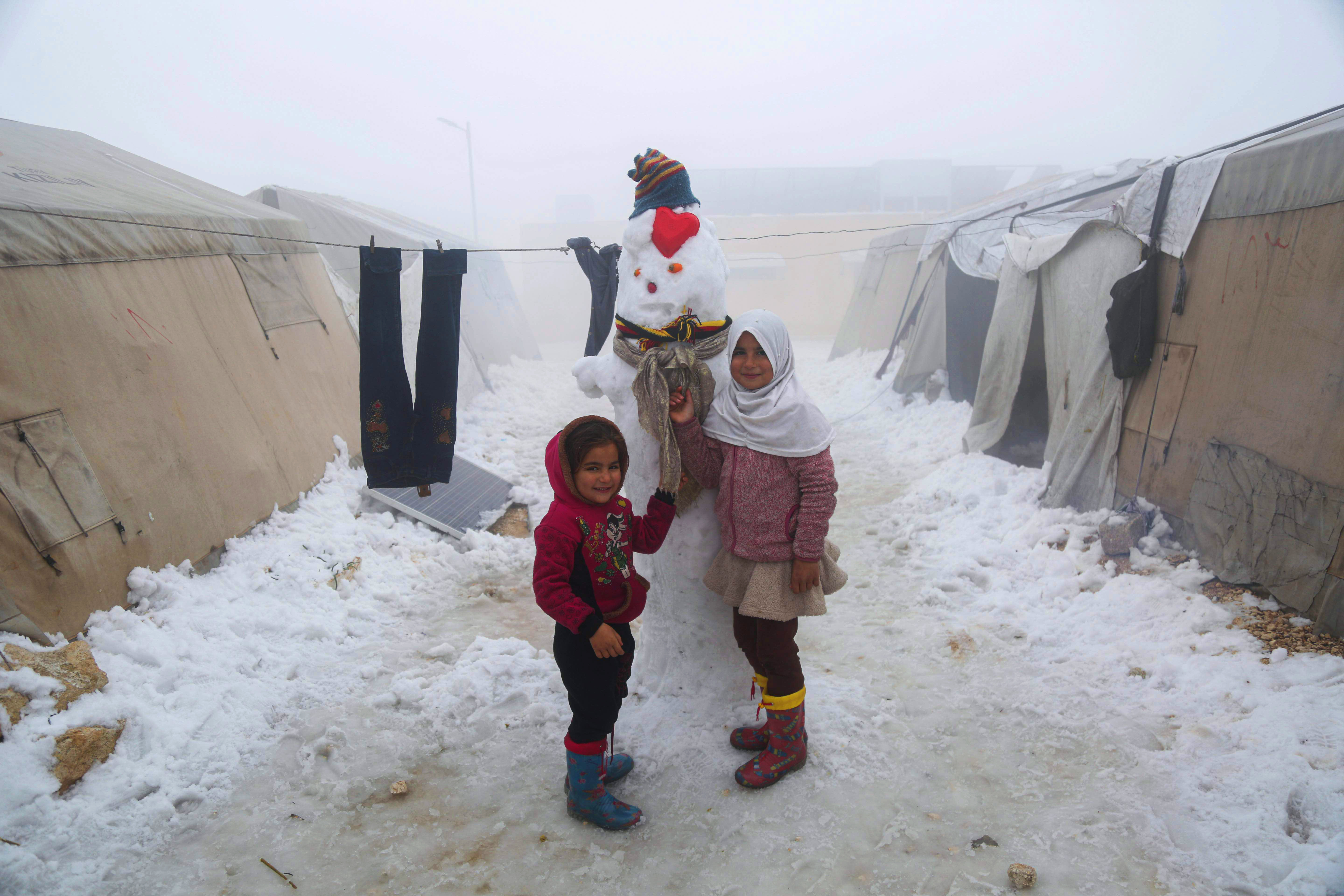 Two girls stand next to a snowman with tents around.
