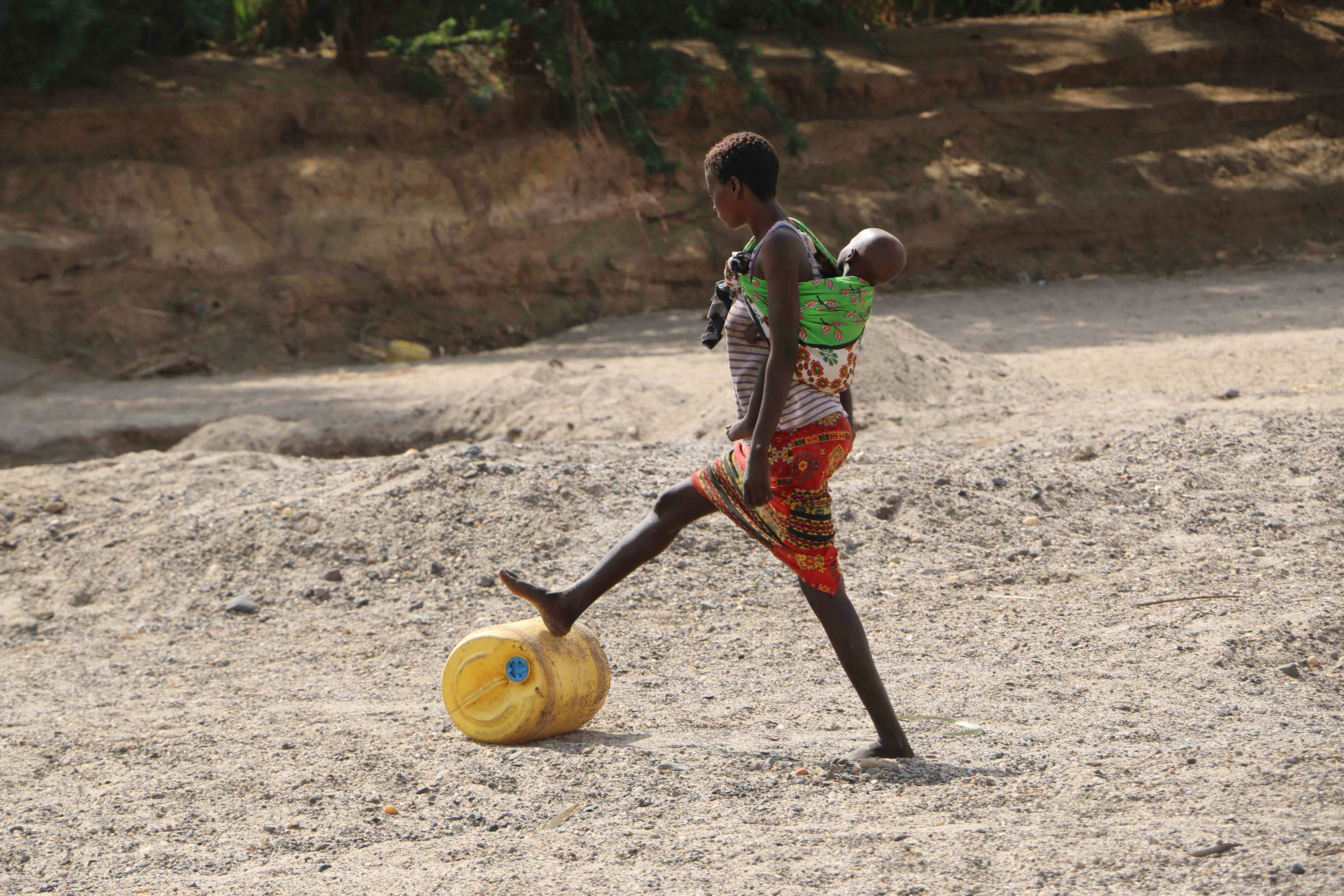 a woman carrying a baby on her back kicks a container of water