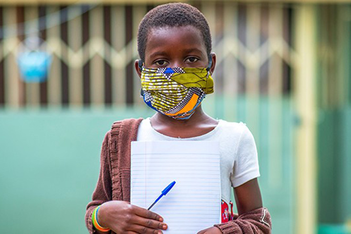 A masked child holds up a piece of paper and a pen.