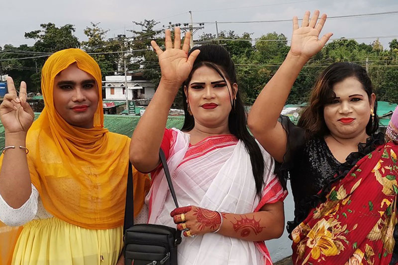 three men from Bangladesh dressed in colorful saris raise their hand to the air and one of them indicates the sign of victory