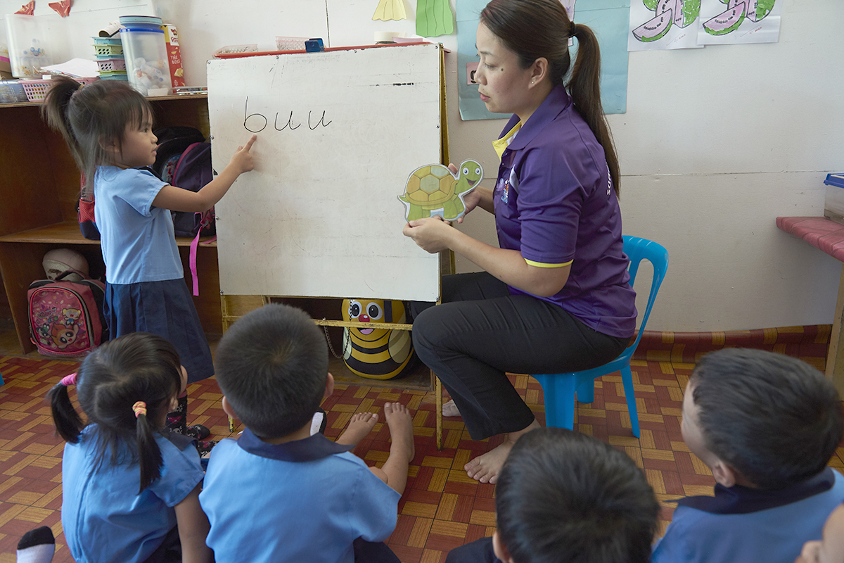 A girl stands in front of the classroom pointing at letters in a notepad next to the teacher.
