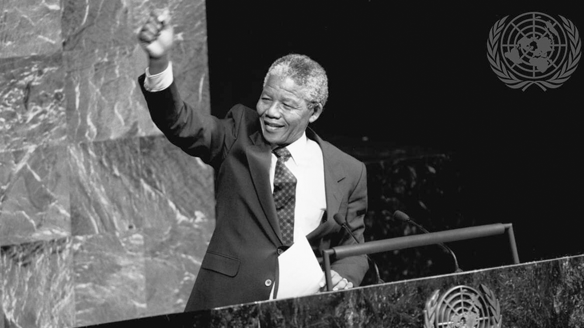 Foto en blanco y negro de Nelson Mandela sonriendo, con el puño en alto en señal de celebración, en el Salón de la Asamblea de las Naciones Unidas