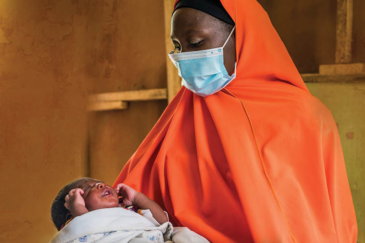 A woman wearing a facemask holds a baby.