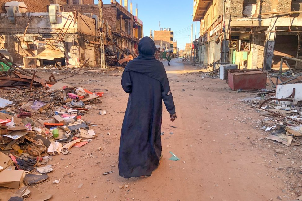 A woman, dressed in black and seen from behind, walks in a street where buildings have been destroyed