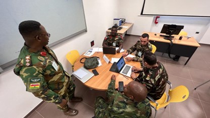 overhead photo of men dressed in military uniforms as they site around a round table looking at a laptop together while one stands up