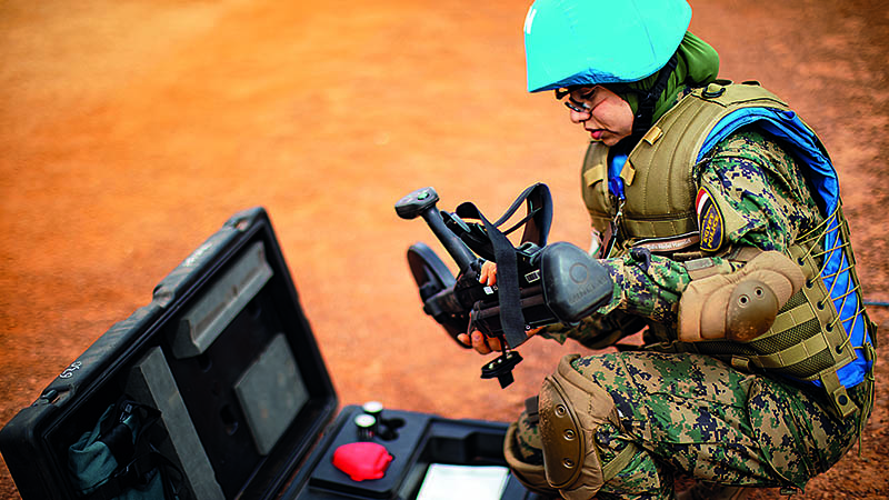 A woman peacekeeper in camouflage clothes and a blue helmet kneels in front of a black suitcase and looks at the black scanning device in her hands.