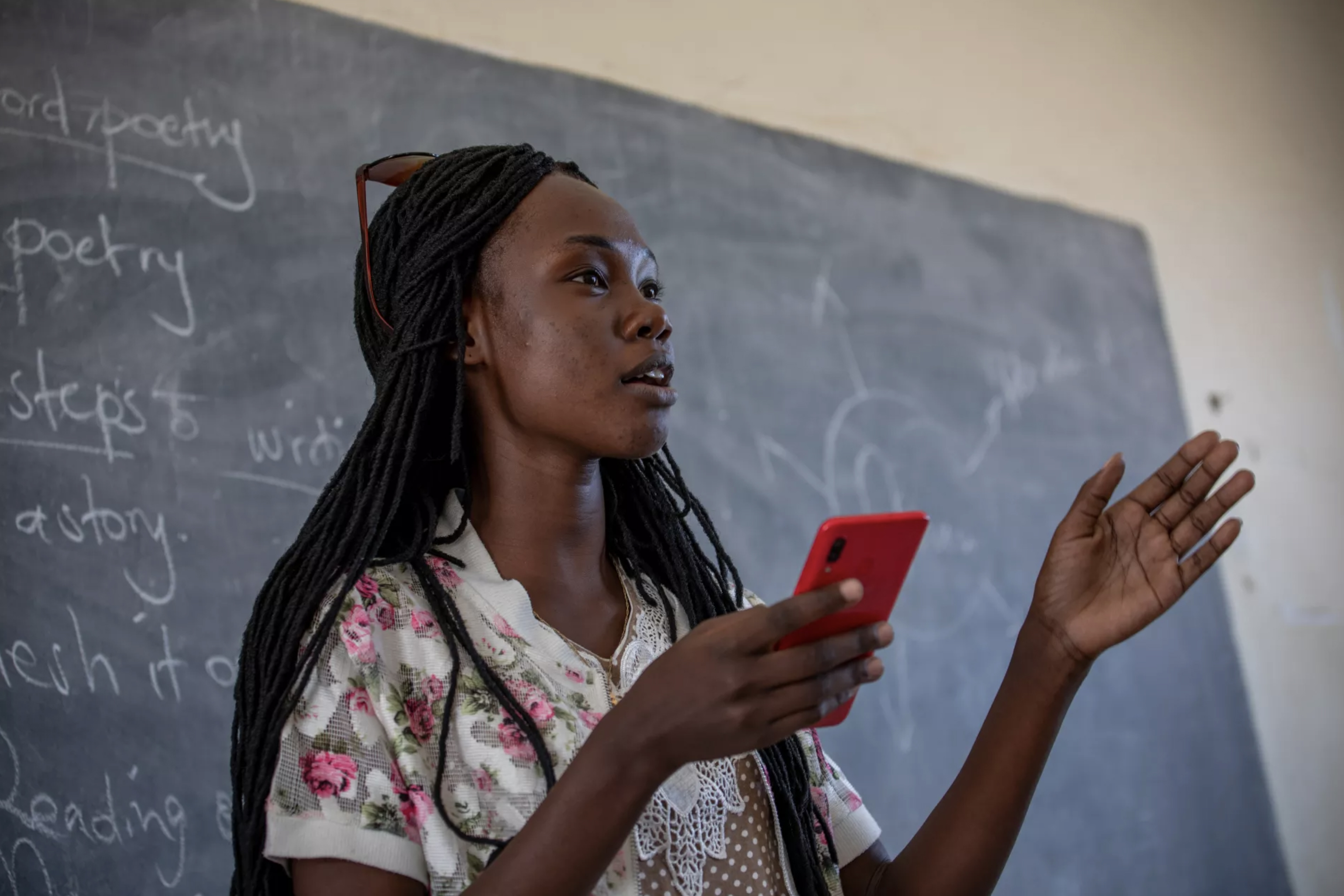 A photograph of a woman standing in front of a chalkboard, gesturing with one hand with a smartphone in the other hand