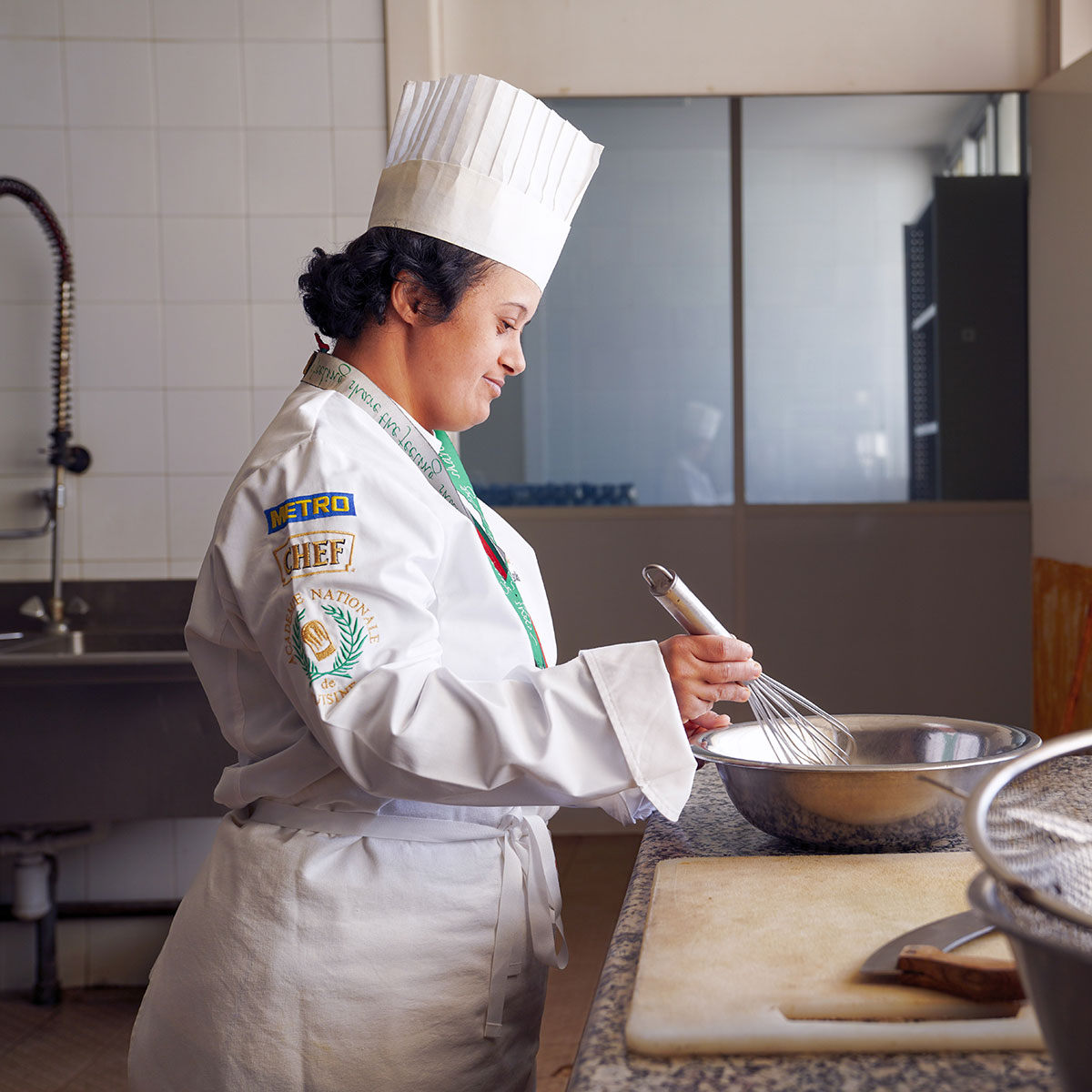 Lobna is in a chef’s hat stands, in a commercial kitchen, smiling down at her mixing bowl and holding a whisk.