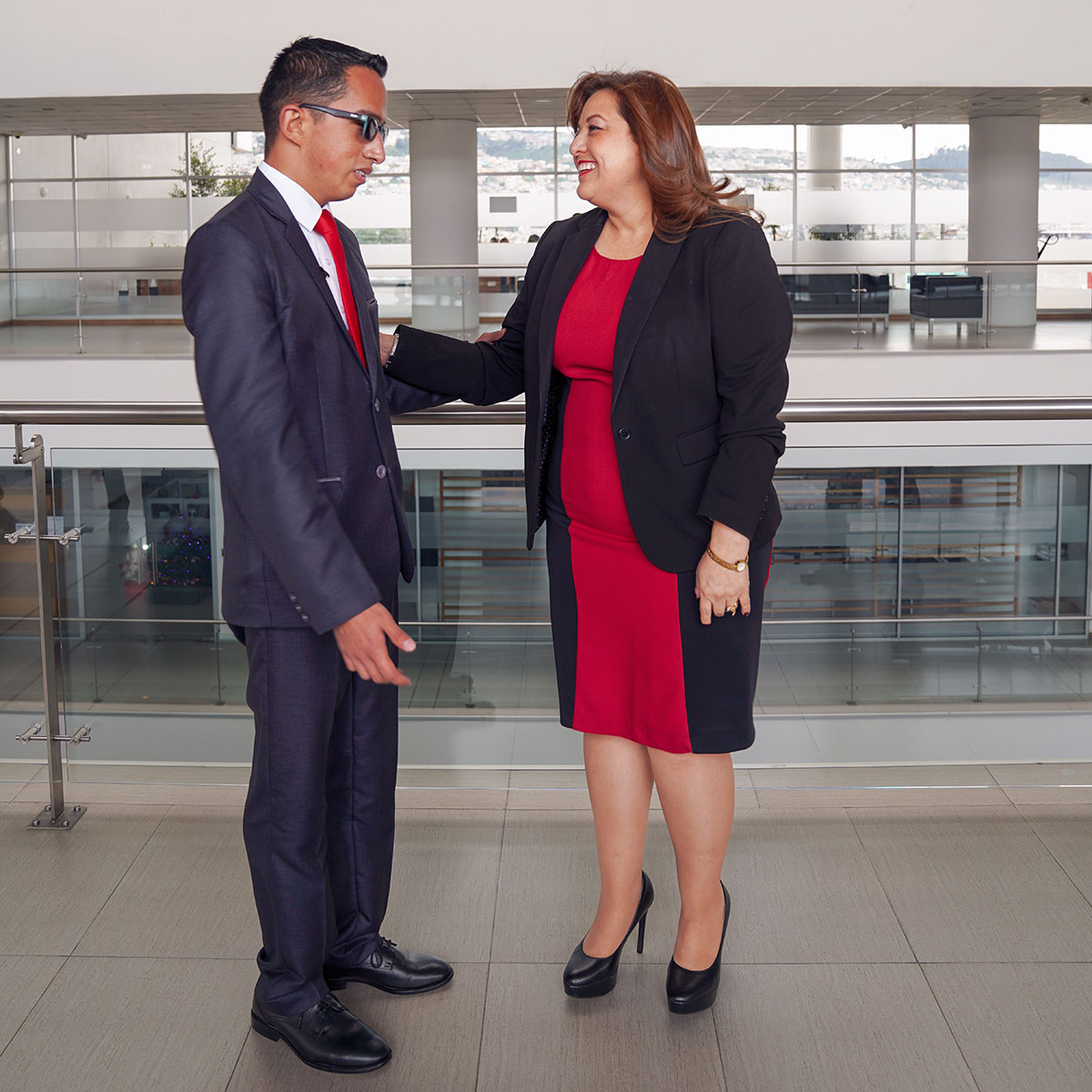 Alex and a woman in business attire stand in a glass office building, facing each other as they converse.