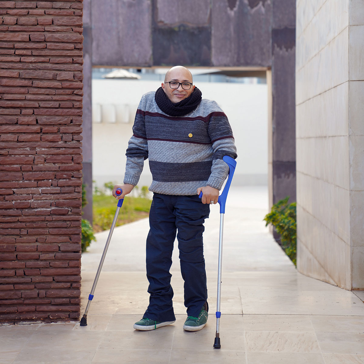 Ahmed wearing a scarf and sweater stands in a courtyard, supporting himself on forearm crutches