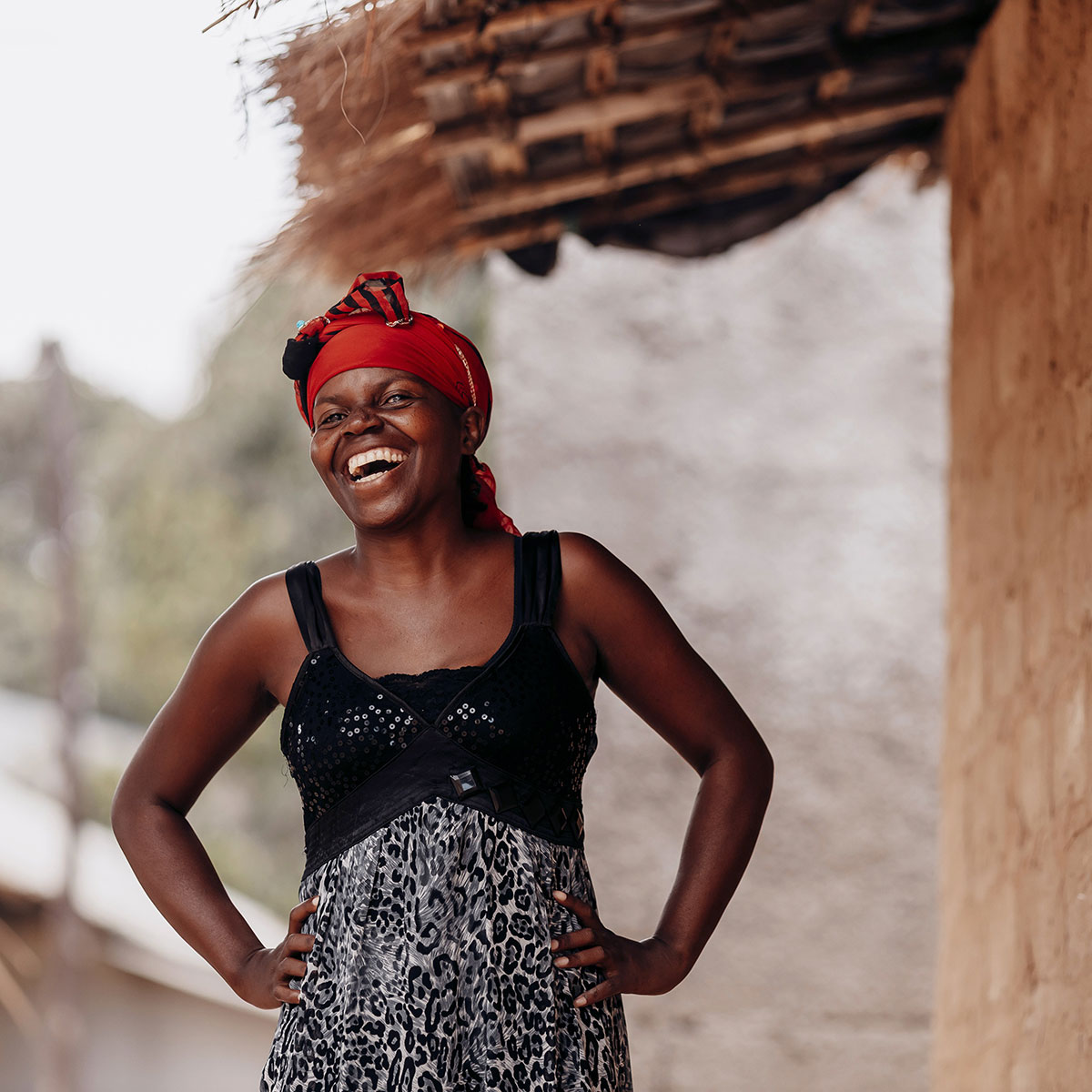 Anchita, laughing, in a leopard-print dress with a black sequined bodice stands next to a mud-brick house, hands on her hips