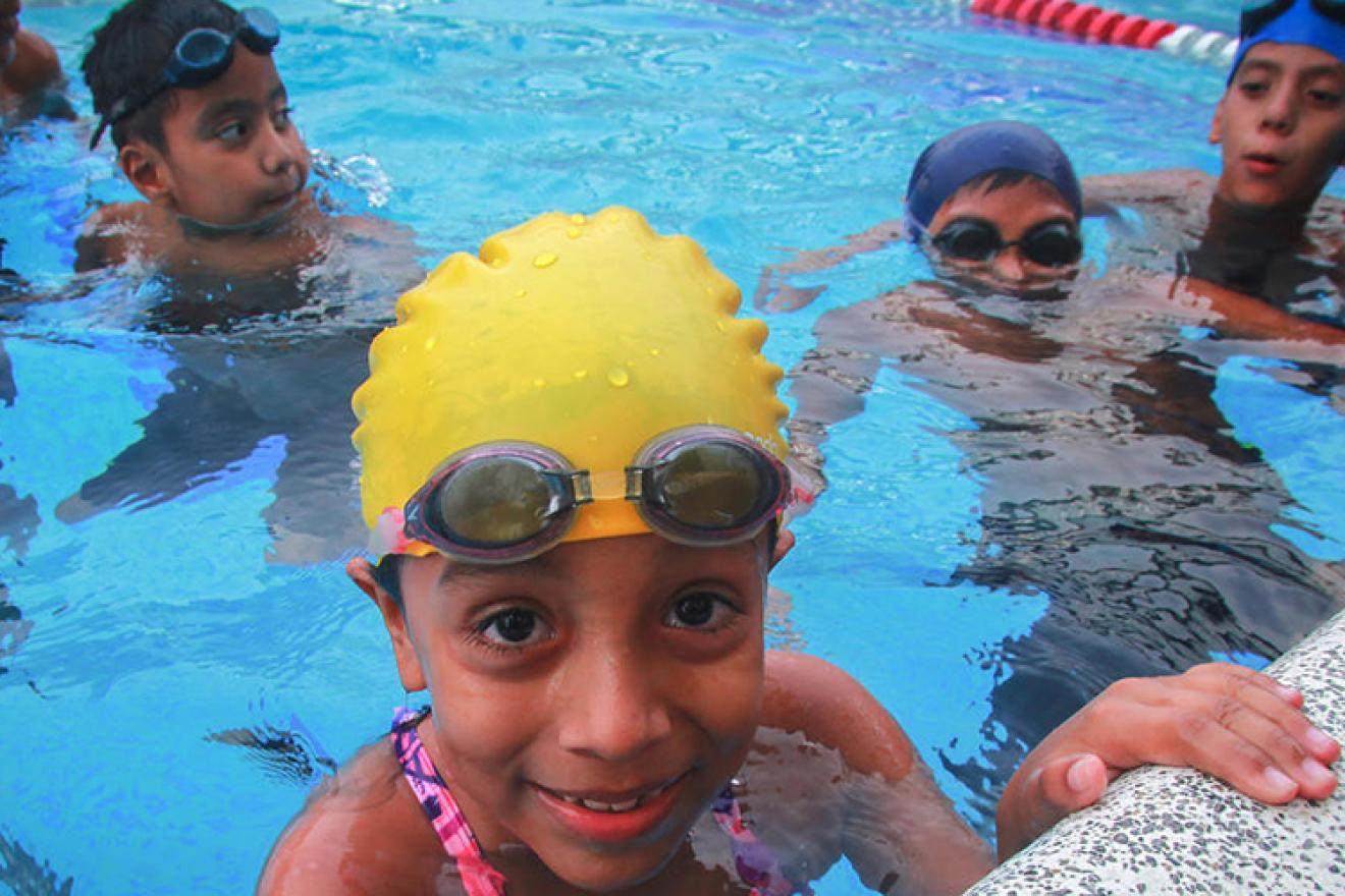 Kids in a swimming pool, with swimming glasses and cap. A girl looks into the camera and smiles