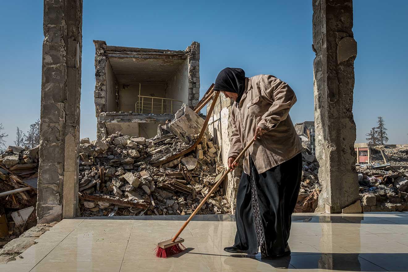 Une femme balaie le sol d'un bâtiment détruit, en face des ruines d'autres bâtiments.