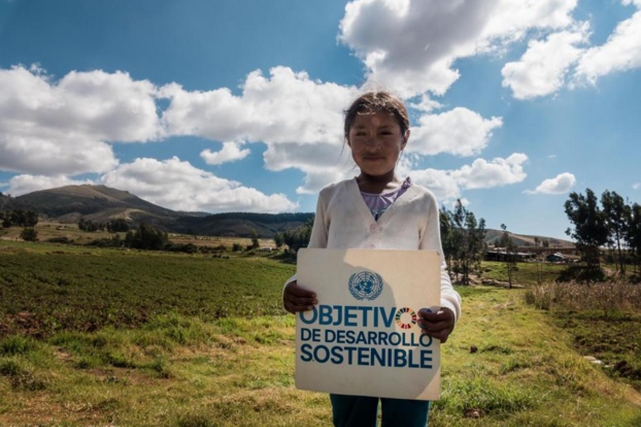 Una niña sostiene un cartel de los Objetivos de Desarrollo Sostenible (ODS) con un paisaje verde de fondo. 